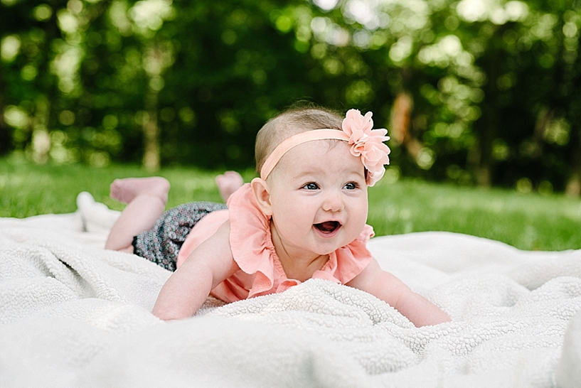 6 month old girl wearing coral shirt with headband laying on blanket in backyard laughing