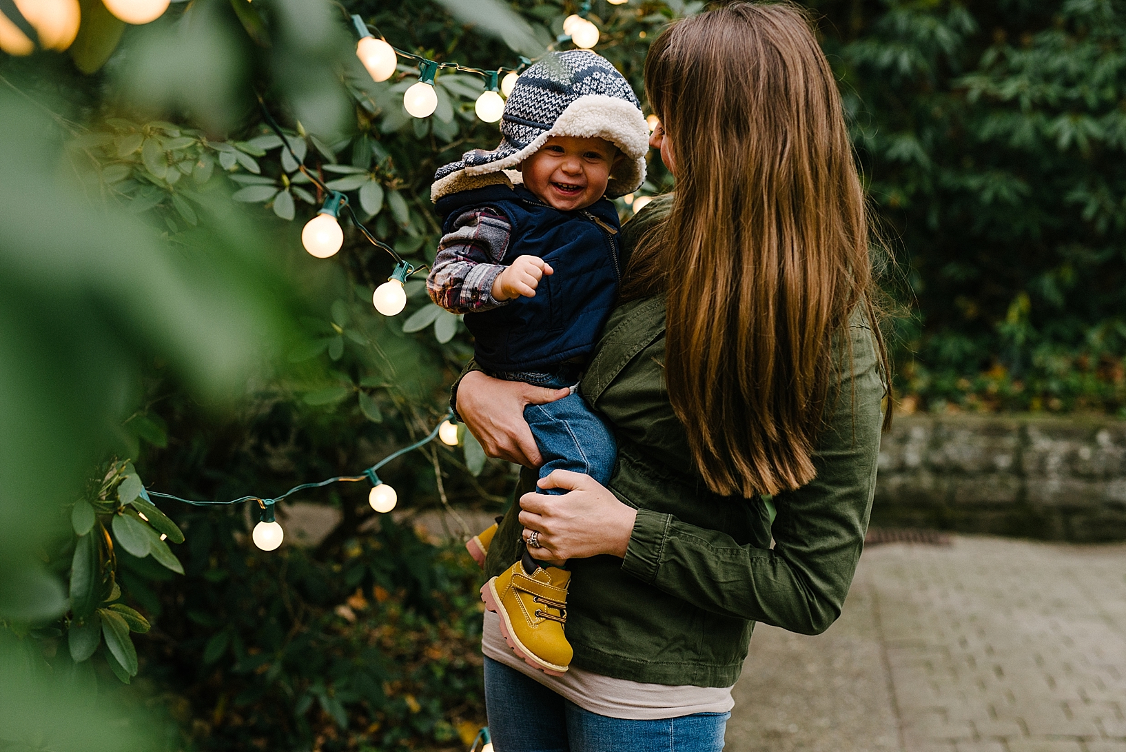 woman holding toddler son wearing trapper hat standing by string lights