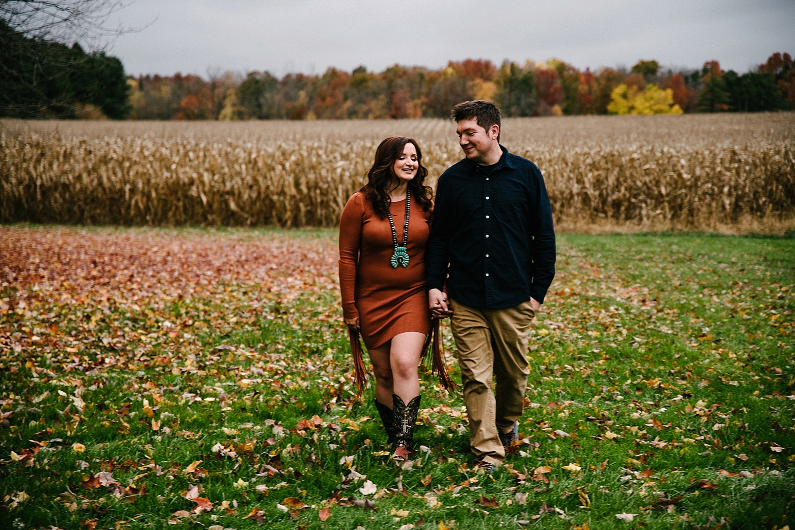 couple walking holding hands with cornfield in the background and leaves on the ground
