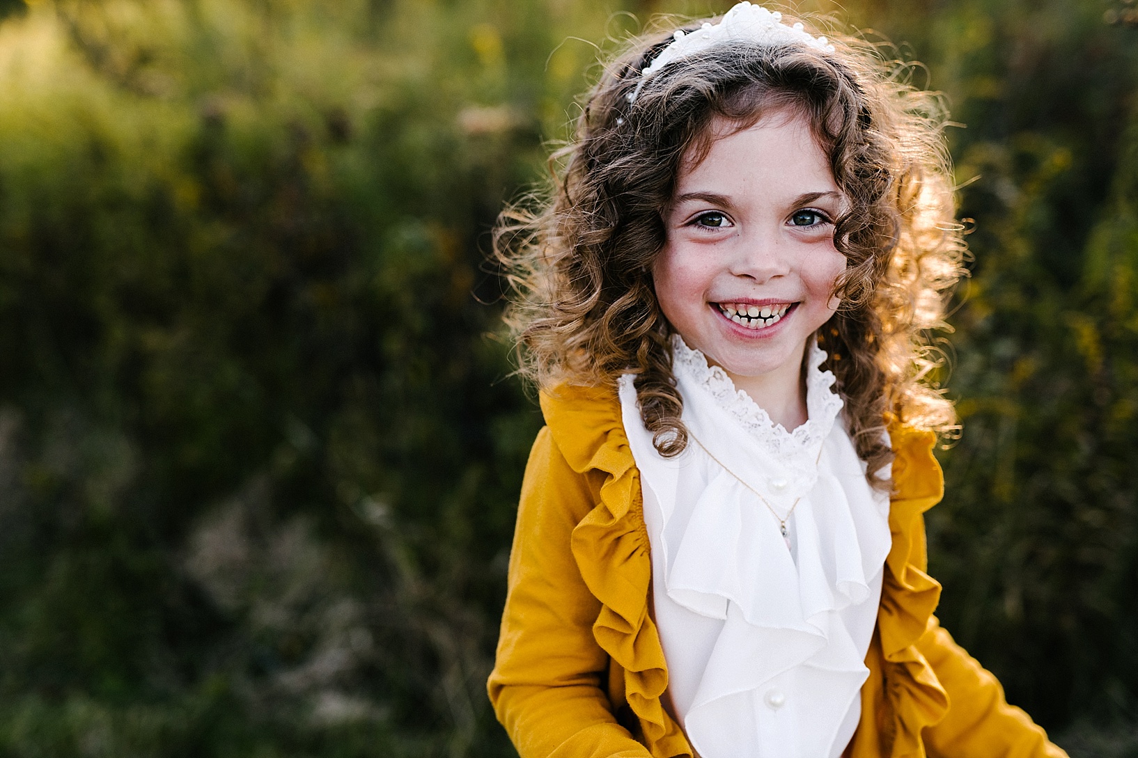 young girl with curly hair wearing mustard yellow sweater smiling at camera