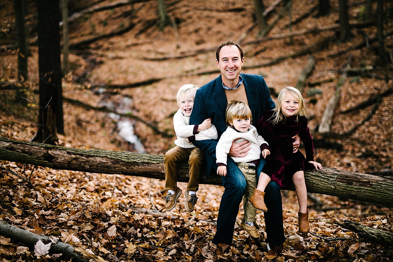 Dad sitting on fallen tree log holding three young kids