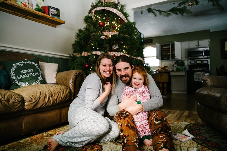 parents with toddler girl sitting in front of christmas tree on christmas morning