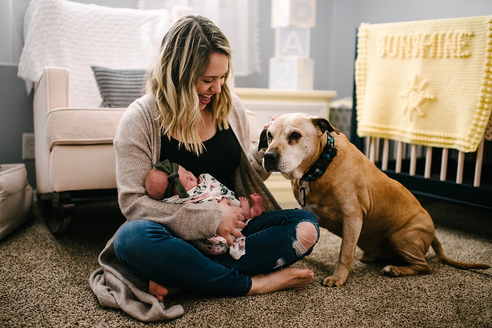 mother holding newborn sitting on floor of nursery with dog next to her