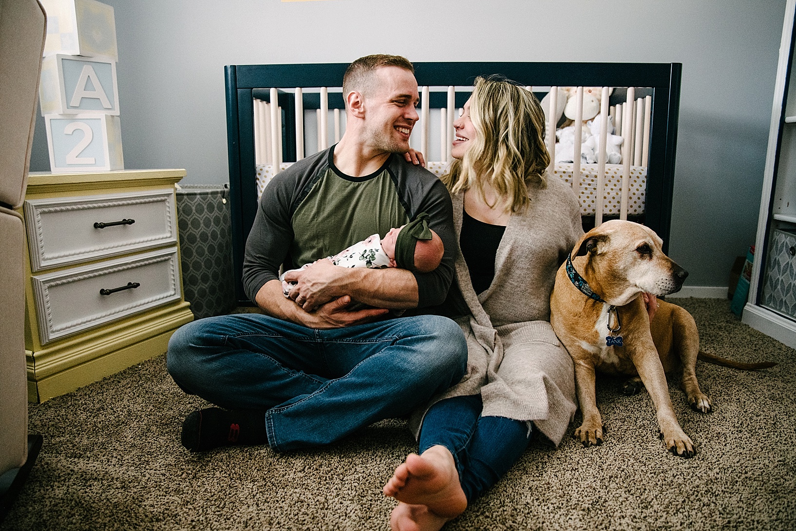 mom and dad sitting on floor of nursery holding newborn with their dog
