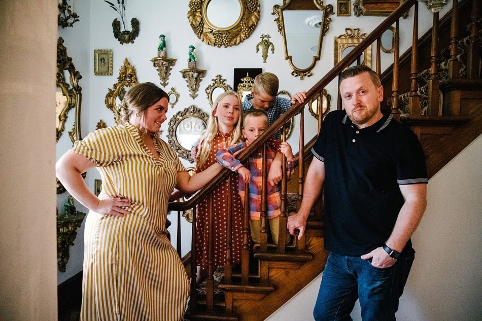Mom, dad, daughter, and two sons stand in front of wooden staircase in quirky family portraits session