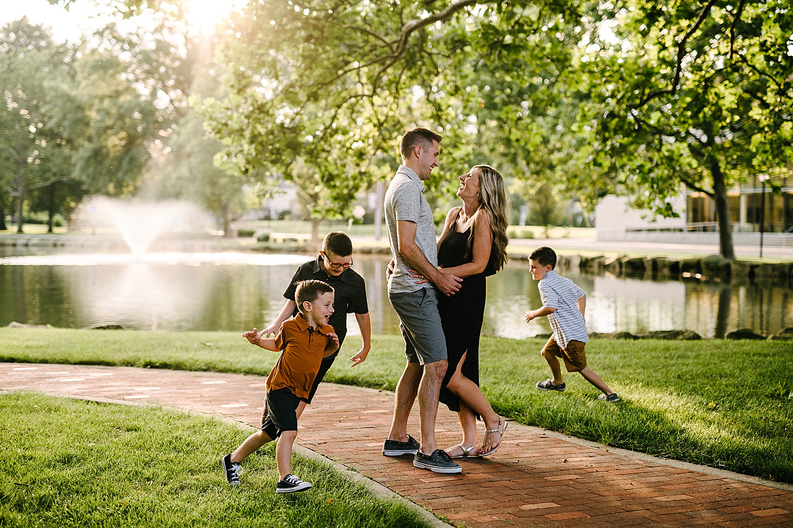 Mom and dad stand smiling and embracing while three boys run in a circle around them in front of Mount Union pond.