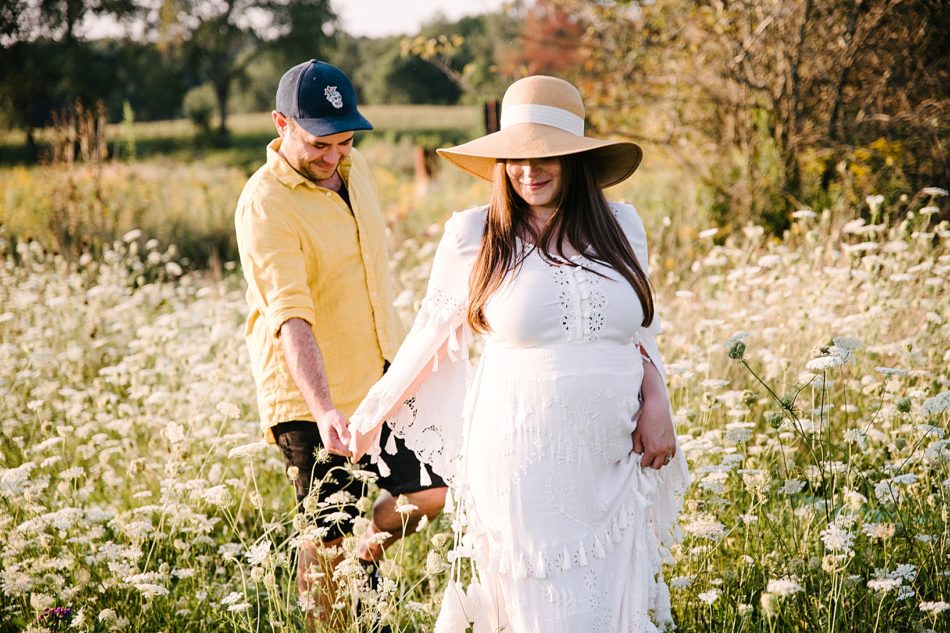 Pregnant woman in white gown holds husbands hand behind her as they walk through a field of white wildflowers on their flower farm.