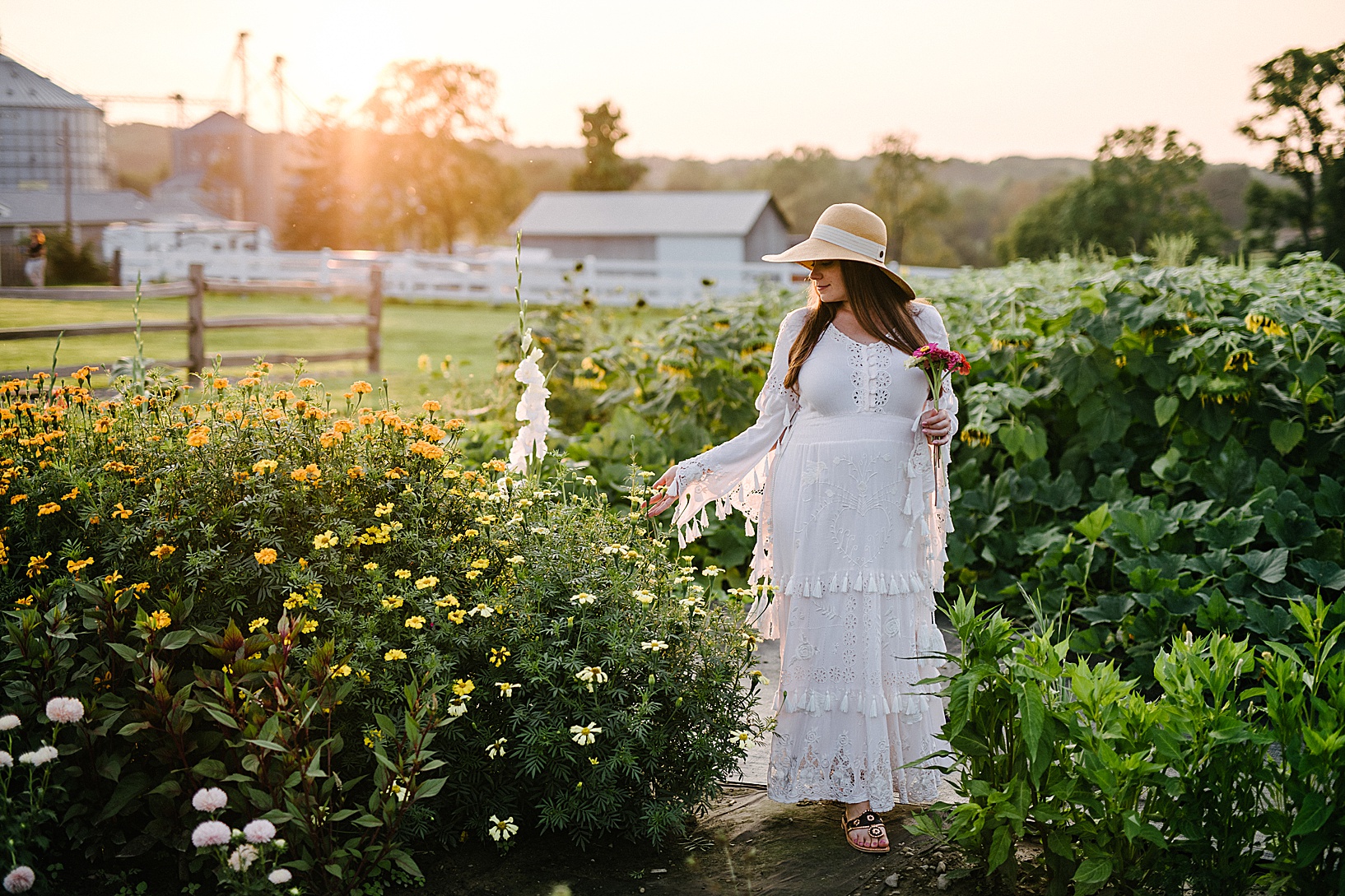Pregnant woman in white Fillyboo gown and sun hat walks through rows of wildflowers