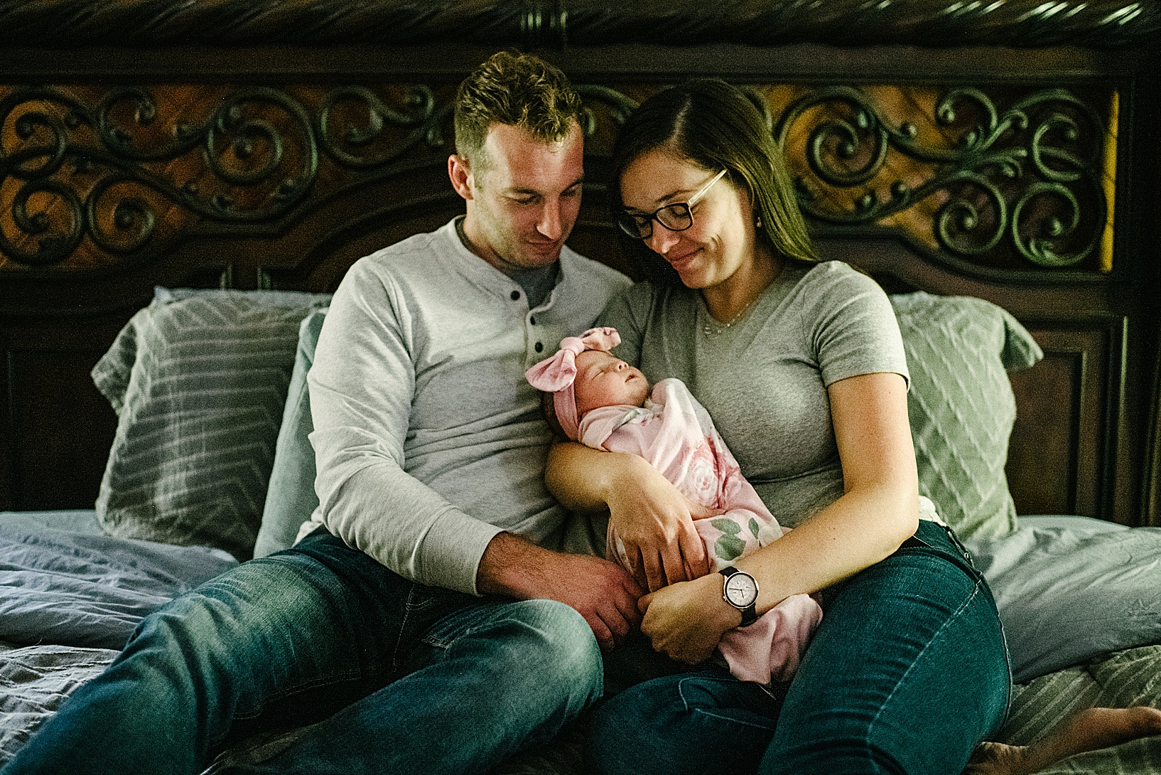 Woman and man cuddle newborn Baby Ava on their bed with beautiful iron headboard in background