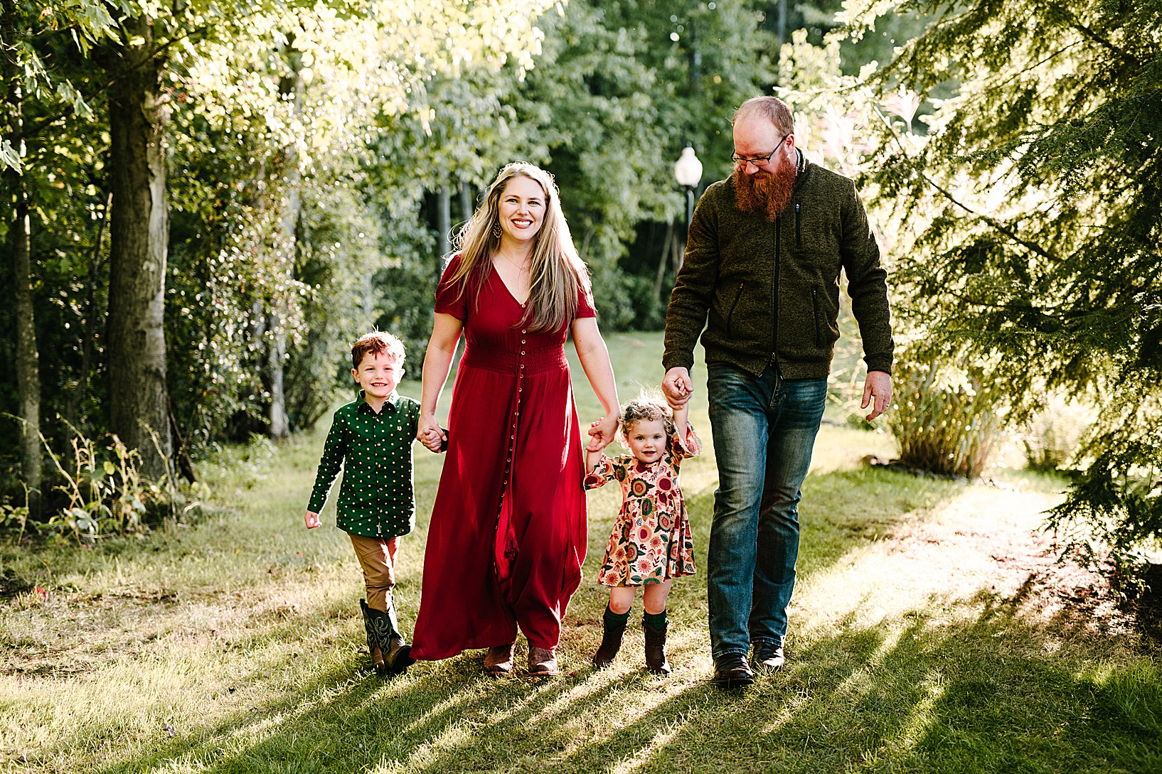 Blonde woman in long red dress and red-bearded man walk through a field while holding their son and daughters hands during this fall family session