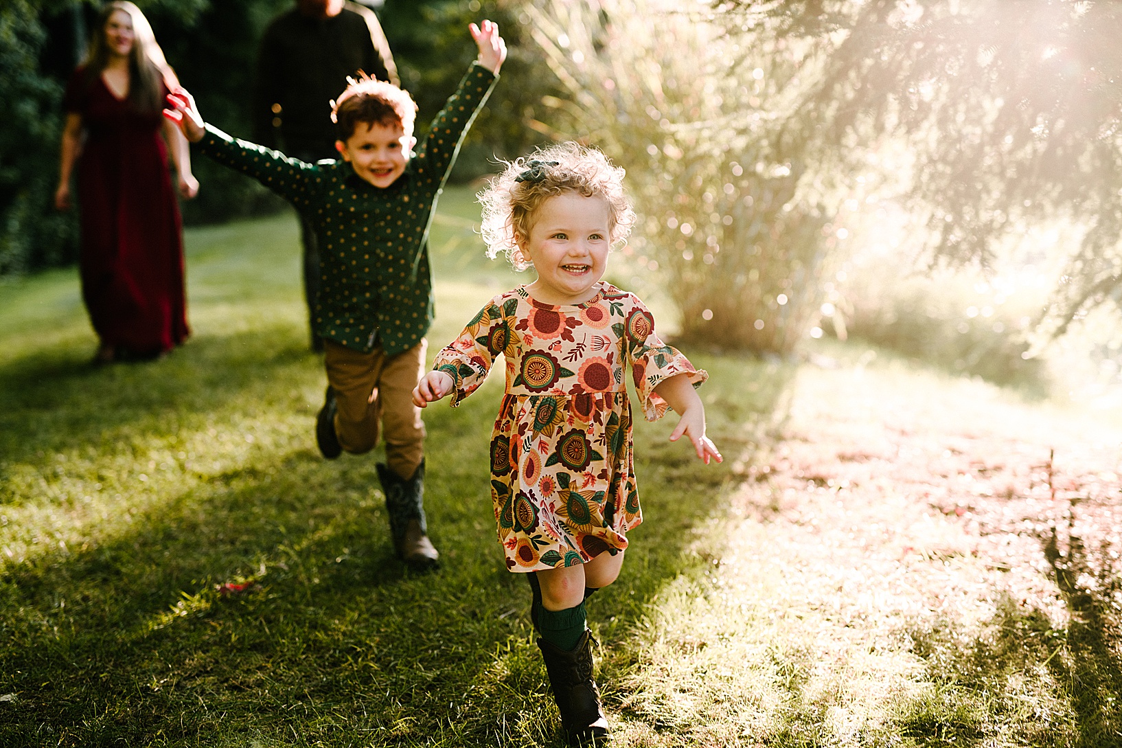 Blonde toddler in fall patterned dress and cowboy boots and her brother in green polka dot shirt and cowboy boots run smiling through a field towards the camera