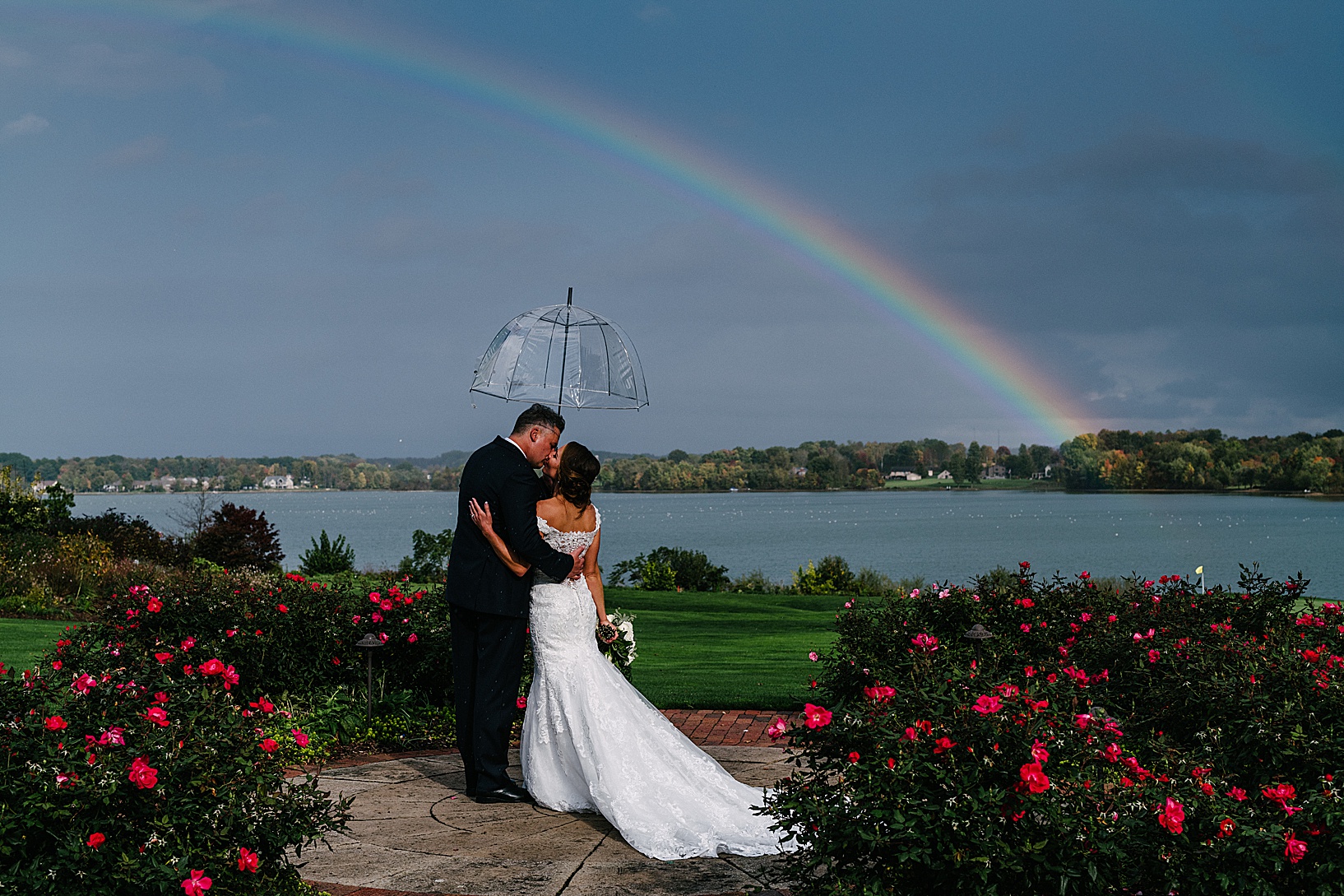 Bride and groom kiss in front of a gorgeous lake holding a clear umbrella and under a rainbow at the Lake Club.