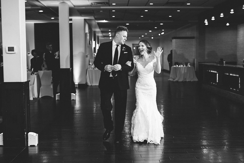 Bride smiles and waves while holding onto grooms arm as they walk into their reception.