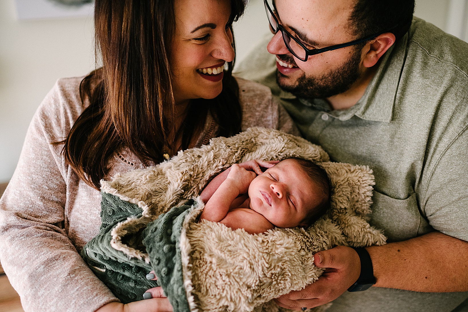 Woman and man smile at each other while holding newborn baby Karlo wrapped in a green fur blanket.