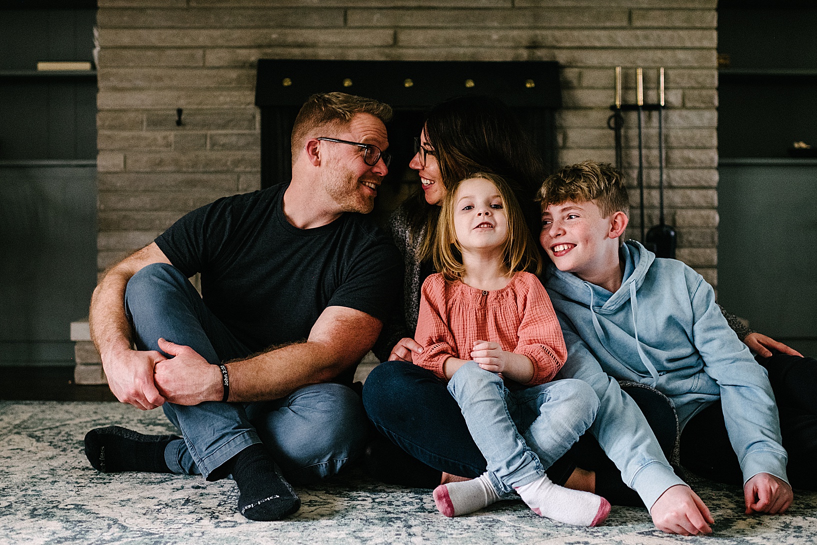 Family of four sits on the floor in front of fireplace with crossed legs and smile at each other during spring lifestyle shoot.