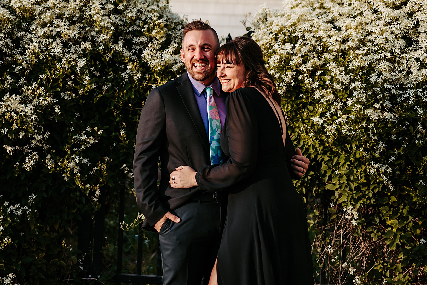 man and woman in formal attire hugging each other and laughing in front of flowers