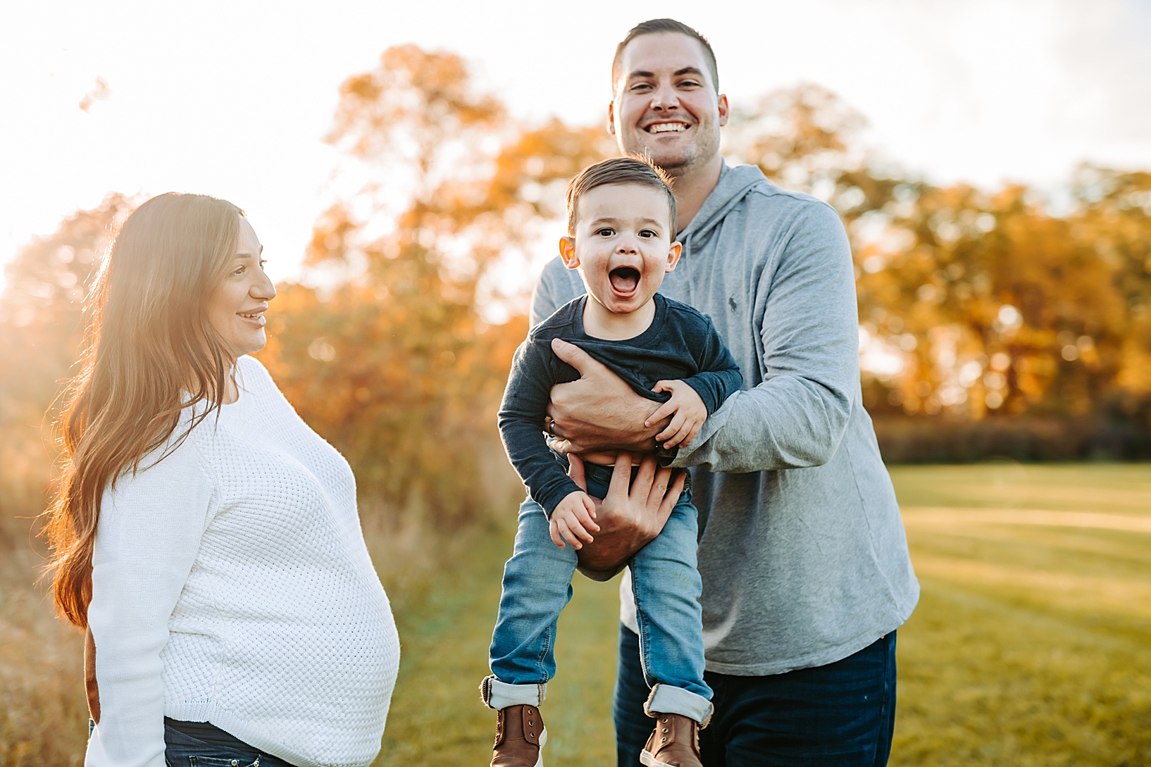 dad swinging laughing little boy in the air while pregnant woman smiles at them