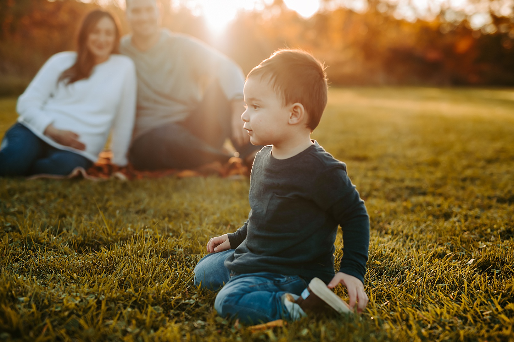 toddler boy sitting in grass with his mom and dad sitting on blanket in the background