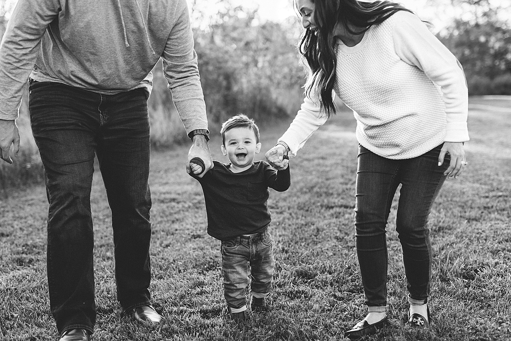 mom and dad holding hands of young toddler boy and walking through field