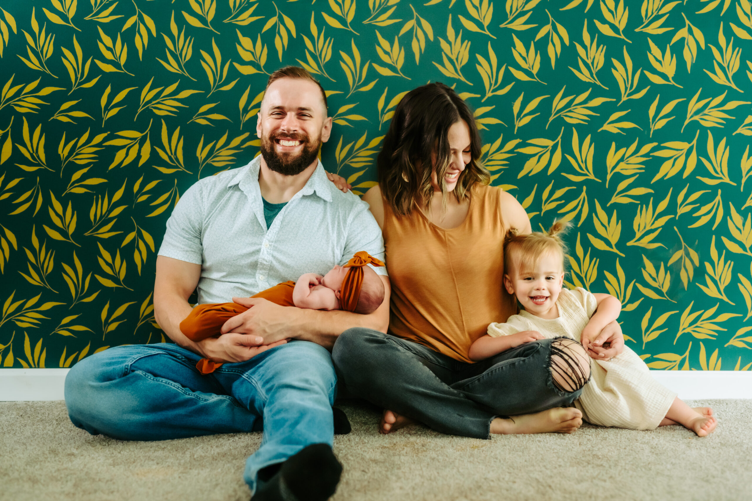 man and woman sitting against green wall with toddler daughter and newborn baby