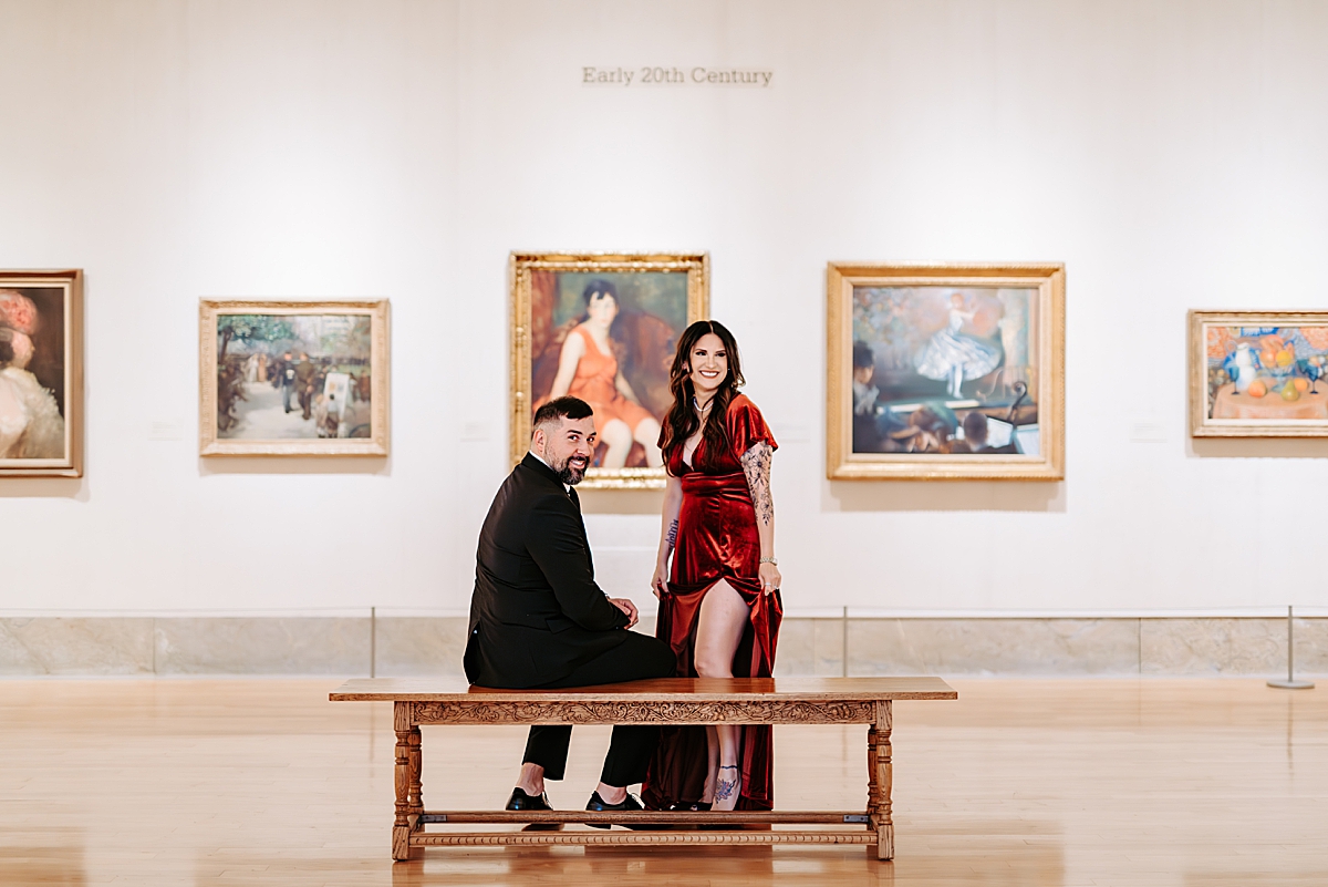 man in suit sitting on bench and woman in red velvet dress standing behind bench in gallery room at The Butler Institute of American Art