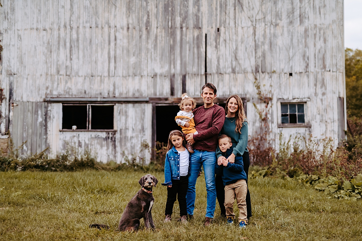 family with young children and dog standing in front of old white barn