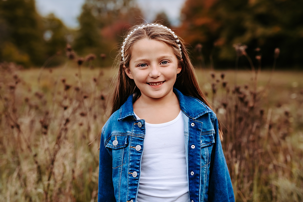 young girl wearing jean jacket and pearl headband standing in field smiling