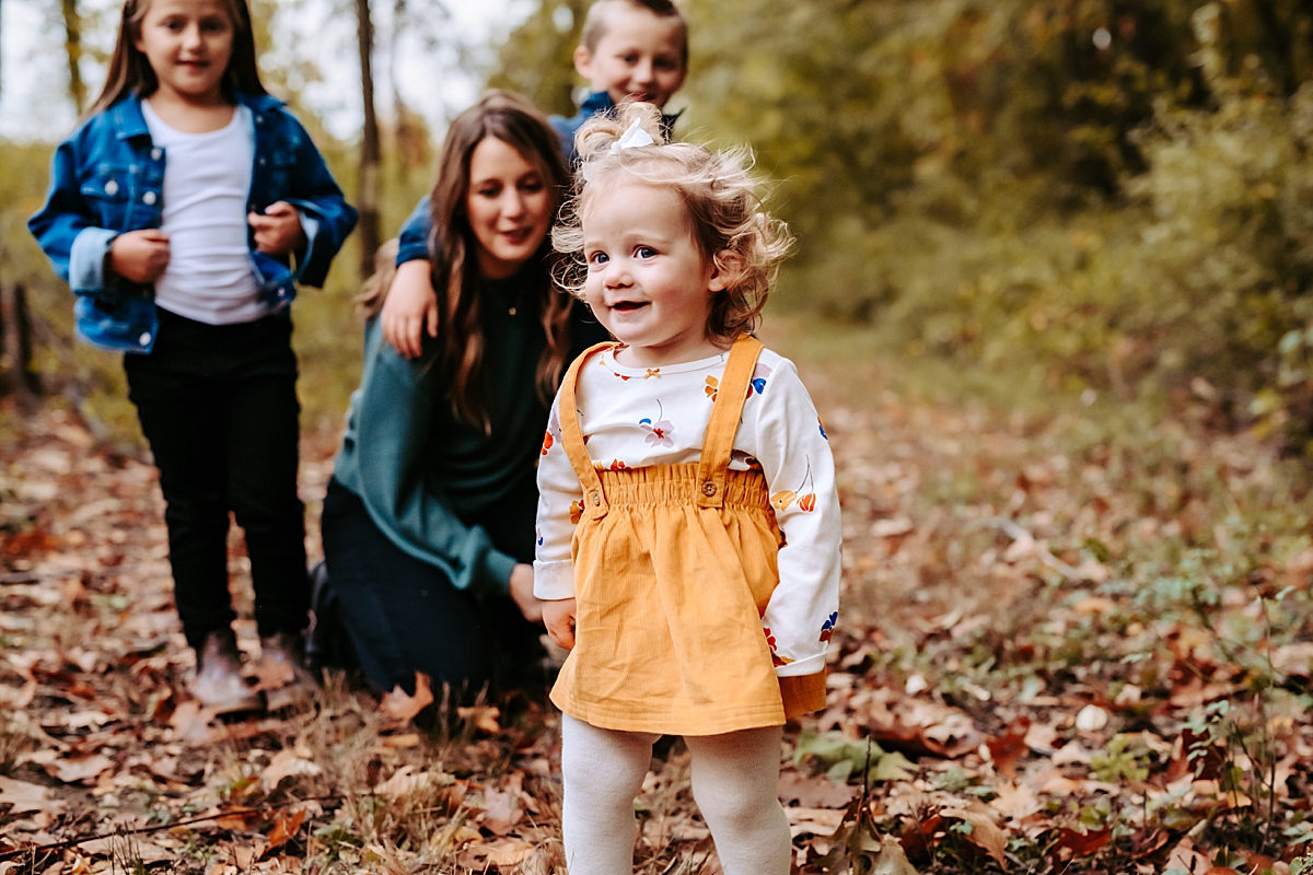 toddler girl smiling standing in front of mom and siblings outdoors in the woods