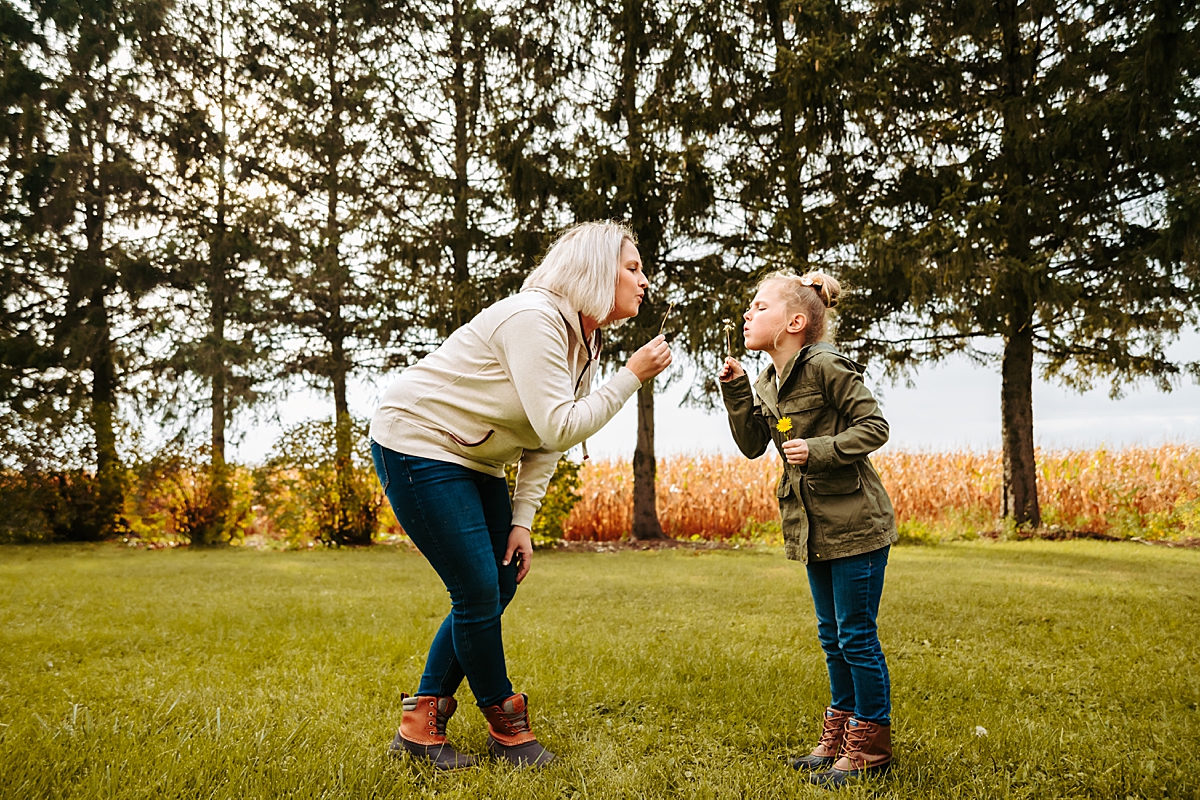 mother and daughter blowing dandelions in front of corn field