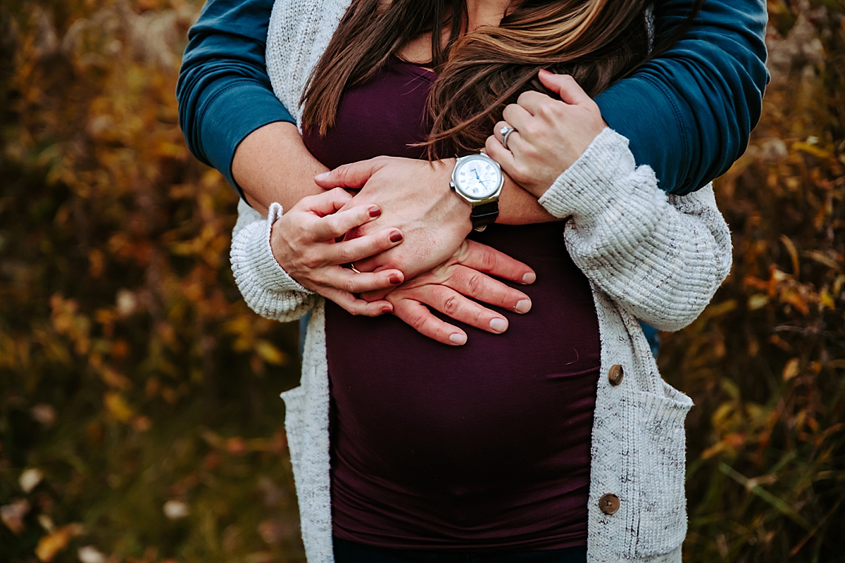 man's arms wrapped around wife's shoulders and hands resting on her pregnant belly