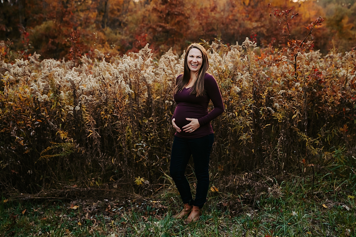 pregnant woman laughing with hands on her belly standing in front of fall field