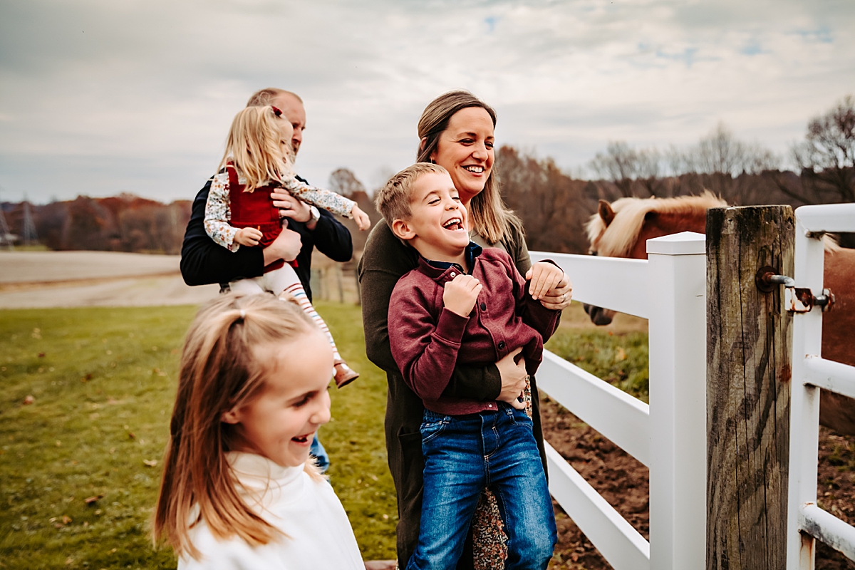 parents with young kids laughing and looking at horses by white fence