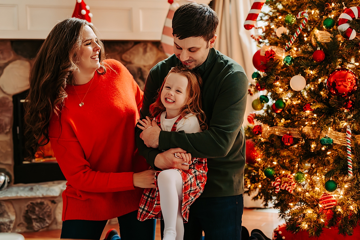 mom and dad with young girl laughing next to Christmas tree and fireplace