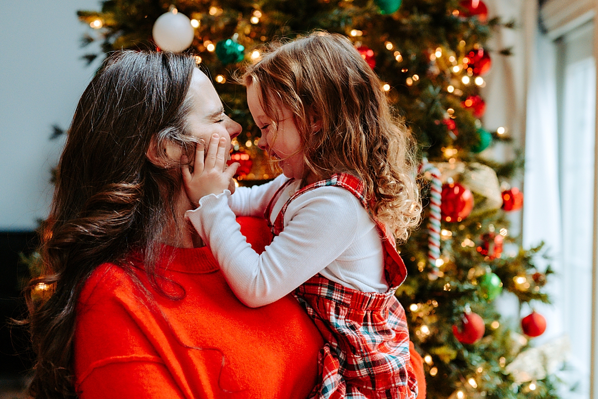 daughter in mom's arms squeezing her cheeks in front of Christmas tree