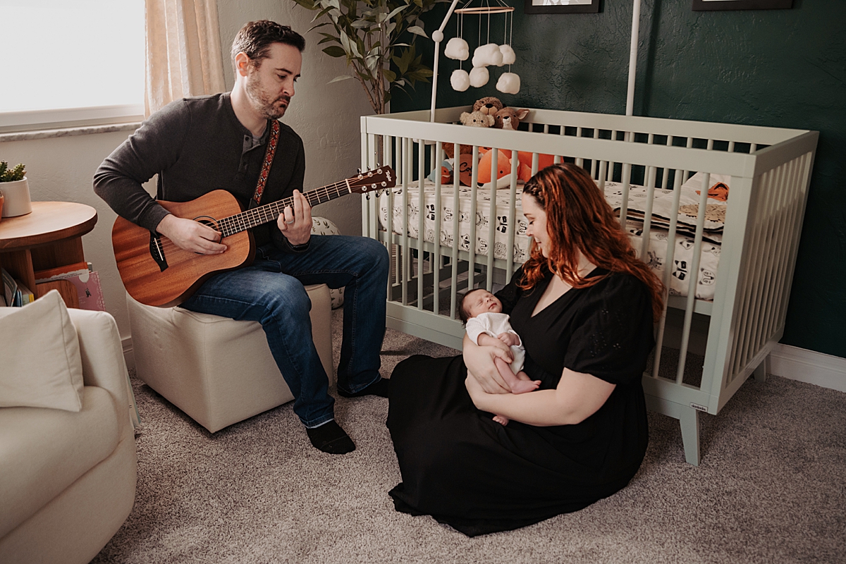 mom holding newborn baby sitting on floor in front of crib while dad plays the guitar