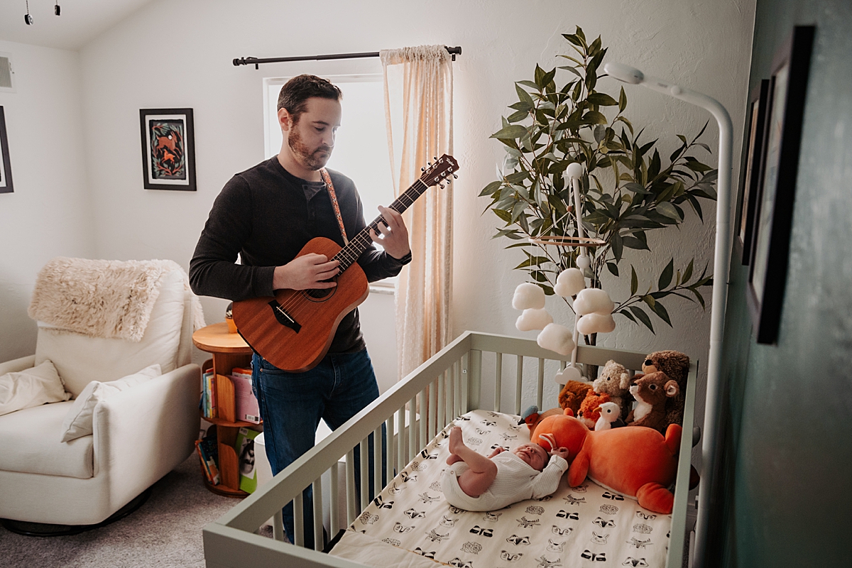 dad playing acoustic guitar standing in front of crib with newborn in it