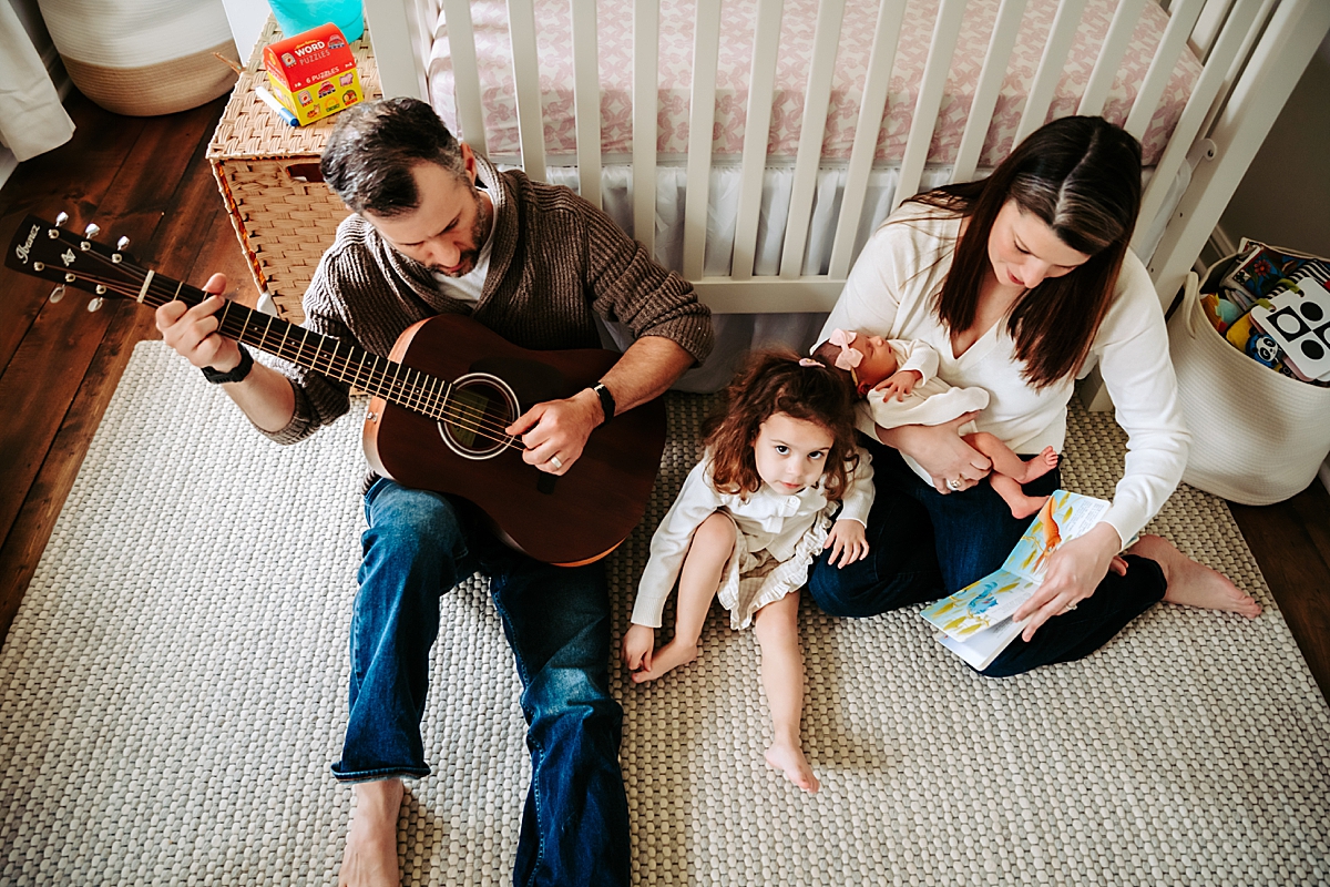 dad playing guitar sitting in front of crib with toddler and mom holding newborn
