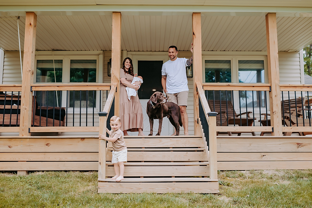 A family stands on the front porch of a house. A woman holding a baby stands to the left, while a man with a dog is to the right during their newborn session. A young child in a tan outfit stands on the steps. The porch features wooden railings and cozy outdoor furniture.