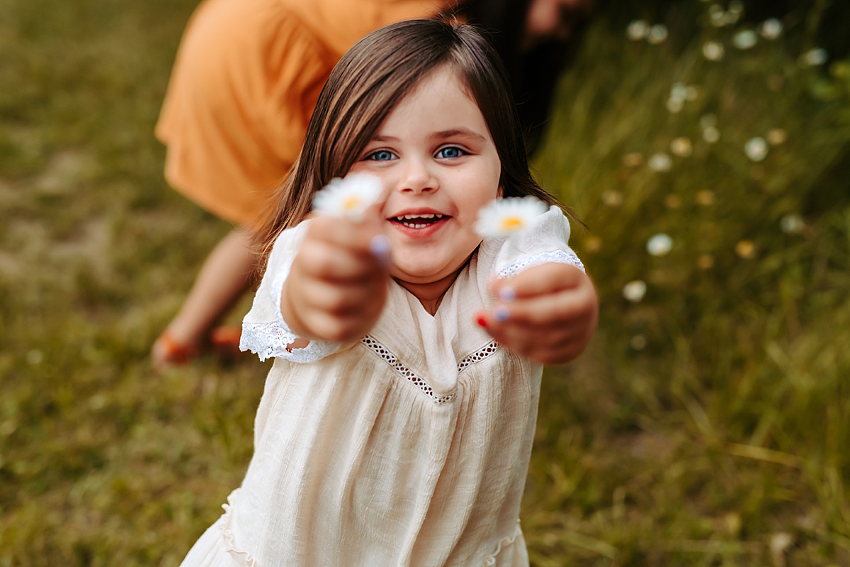 A young child with long brown hair and blue eyes smiles widely while holding two small daisies toward the camera.She is wearing a white dress with lace details. The background is grassy, capturing the essence of summer, with another person partially visible in the distance.