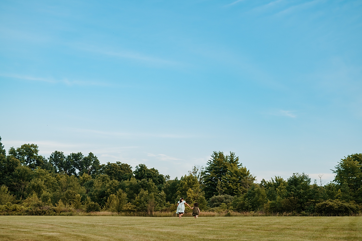 Two young girls in linen dresses hold hands as they walk away across a vast, grassy field towards a line of trees under the clear blue summer sky. Shot by Carlyn K Photography.