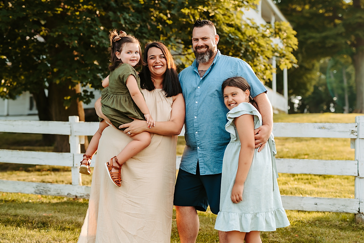 A family of four stands outside in front of a white fence. The mother holds a young girl in her arms while the father embraces their older daughter. They all smile warmly at the camera, with an old farmhouse as their backdrop.