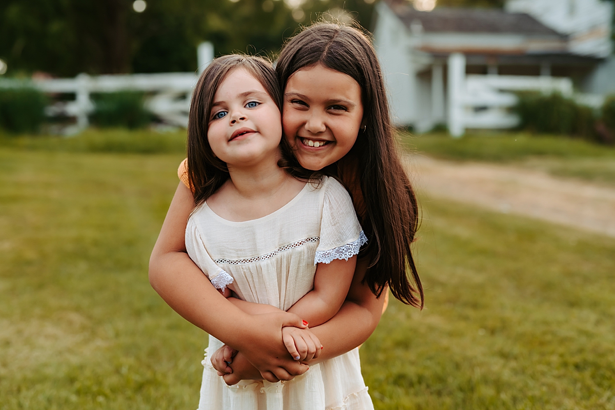 Two young girls with long dark hair are smiling and embracing each other outdoors on a warm summer day. The older girl, standing behind, has her arms wrapped around the younger girl, who stands in front with a light-colored dress. A white fence and a building are visible in the background.