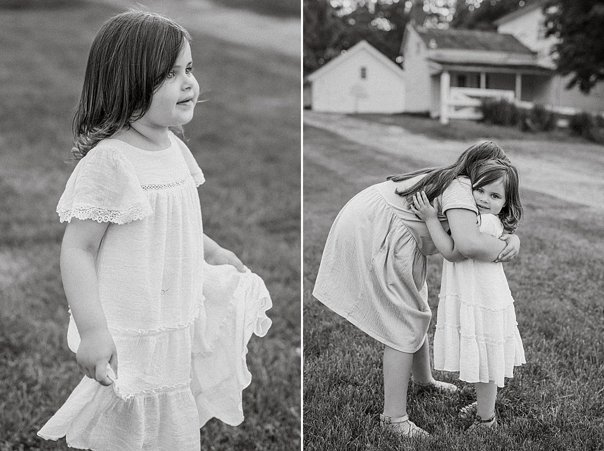 Black and white photo of two young girls in a grassy outdoor setting. On the left, a girl in a white dress stands alone, looking off to the side. On the right, another girl, also in a white dress, hugs the first girl warmly, with a summer house and trees in the background.