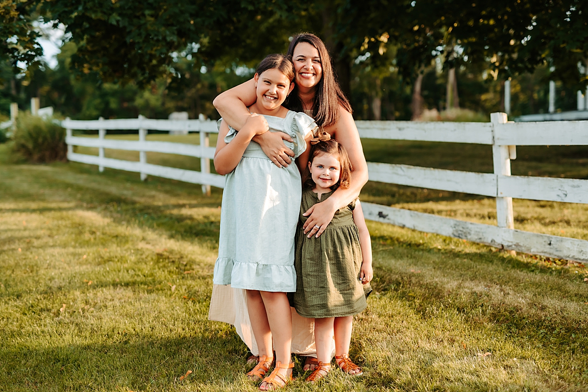 A woman stands outdoors in front of a white fence, smiling and hugging two young girls. The older girl wears a light blue dress and the younger girl wears an olive green dress. The summer sun is shining, and the background is lush and green with trees.