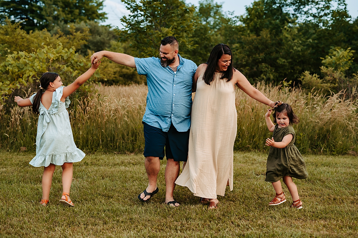 A family of four, consisting of two adults and two children, are holding hands in an outdoor grassy area on a warm summer day. They are smiling and appear joyful, with the adults in the center and a child on either side. Trees and shrubs form the backdrop under a clear blue sky.