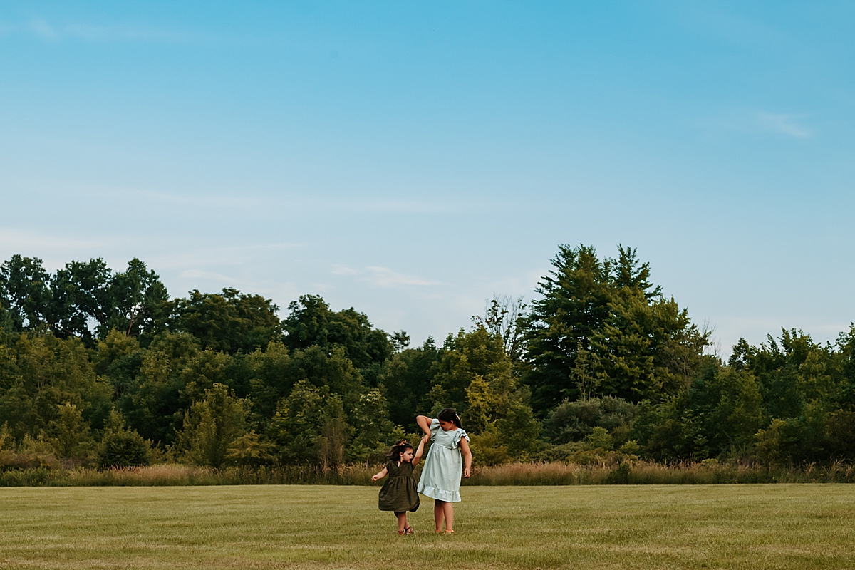 Two children are standing on a lush green field with a backdrop of dense trees under a clear summer sky. Holding hands, they appear to be dancing or playing together. The scene is serene, with the children centrally framed against the expansive natural landscape.