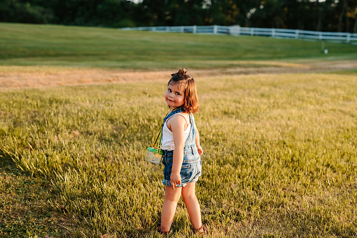 A young girl with short brown hair wearing denim overalls and a white tank top stands in a grassy field, looking back over her shoulder and smiling. She is wearing a bug catcher around her neck on a strap. A white fence and trees are visible in the background, capturing the essence of summer.