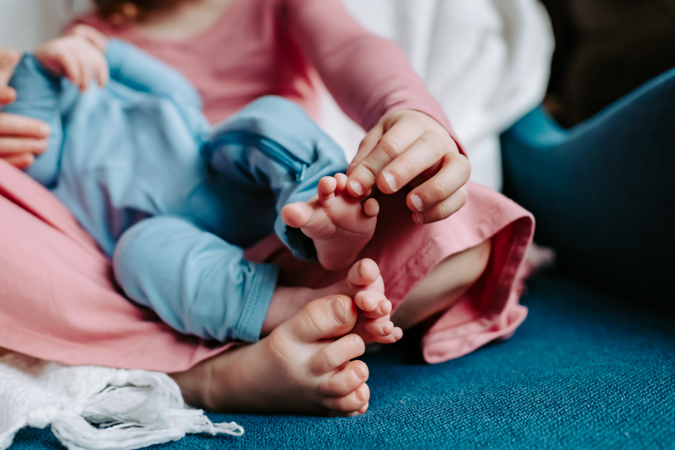 A close-up of a young child in Brecksville gently holding a baby's tiny feet. The baby is dressed in a light blue outfit, while the child is wearing a pink dress. They are sitting on a blue surface with a white blanket partially visible.