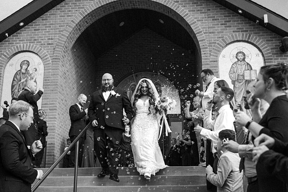 bride and groom exiting down the main steps of Archangel Michael Greek Orthodox Church surrounded by guests blowing bubbles