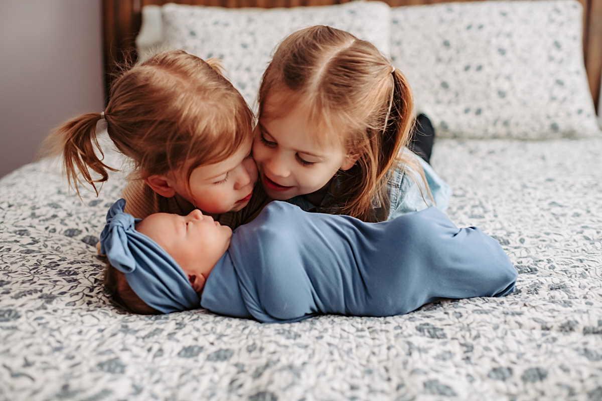 two young girls laying on floral bedspread admiring newborn baby sister in front of them swaddled in blue blanket